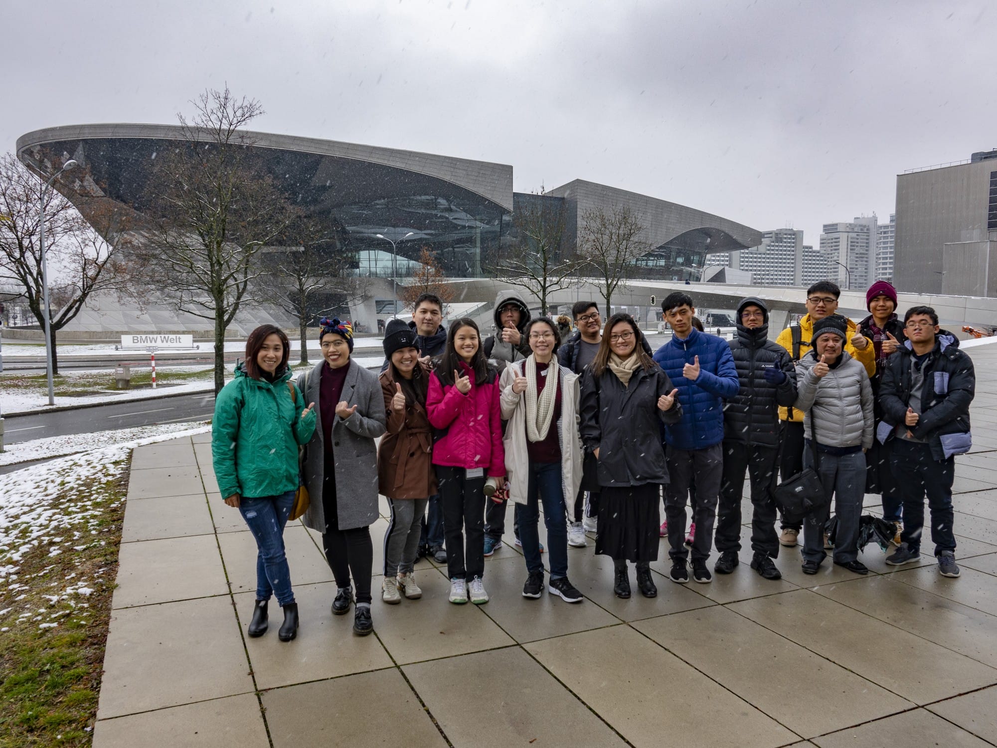 A group of people in winter coats pose with thumbs up outside the BMW Welt building during light snowfall.